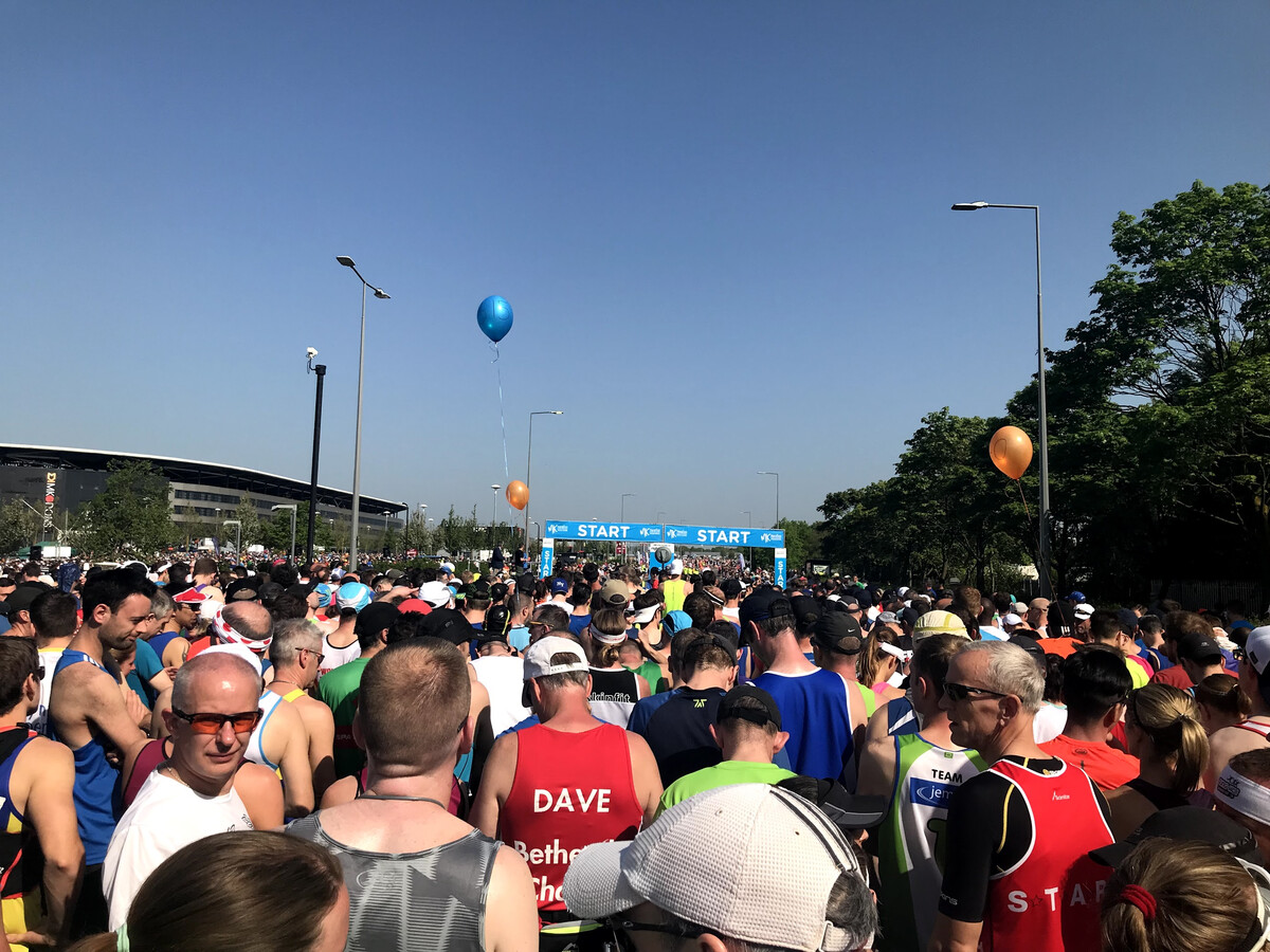 A large crowd of runners packed together at the start line of the MK Marathon, with a blue START gantry and coloured balloons visible above the heads.