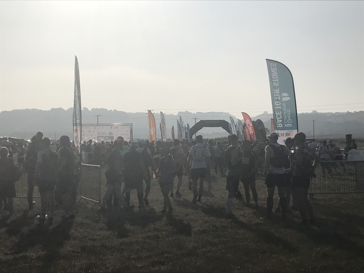 Hundreds of runners and participants mill around a Race to the Stones start and finish area on open grassland at dawn, with the event arch and branded flags visible through the haze.