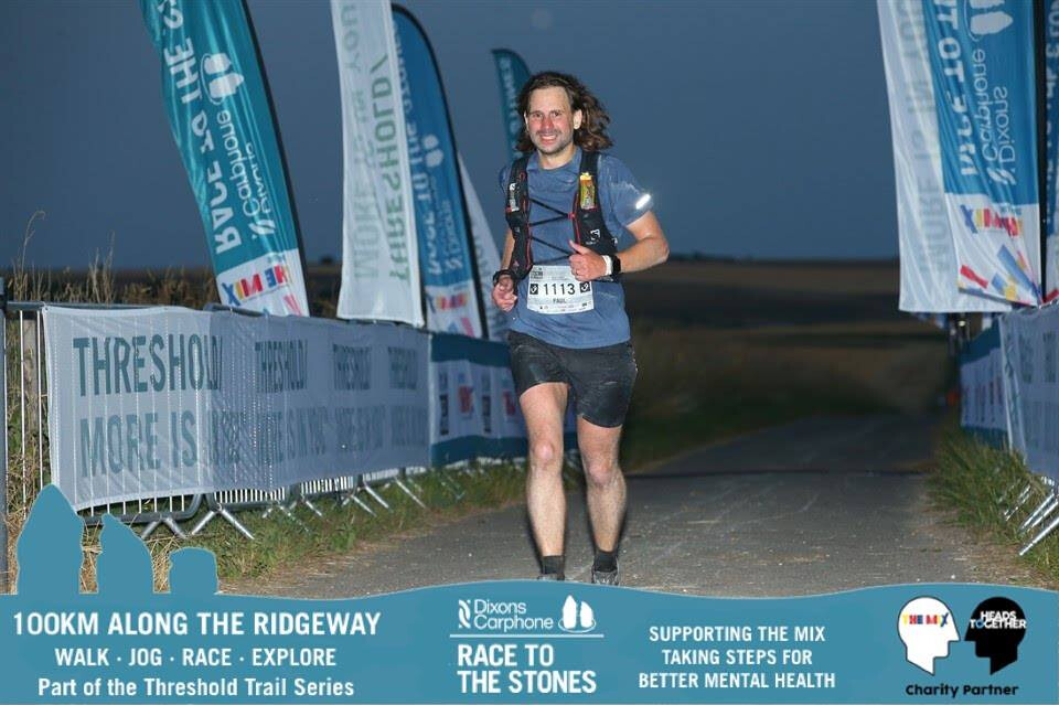 A male runner wearing bib number 1113 runs through an illuminated finish corridor at night during the Race to the Stones 100km event, smiling at the camera.