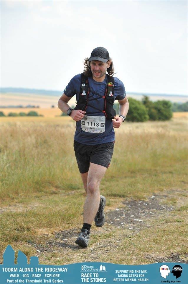A solo male runner wearing bib 1113 and a cap runs across open downland grassland during Race to the Stones, with a wide valley visible behind him.