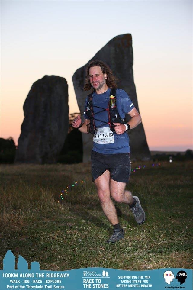 A male runner wearing bib 1113 runs past large standing stones at Avebury at dusk, the sky showing a pink and purple sunset behind the megaliths.