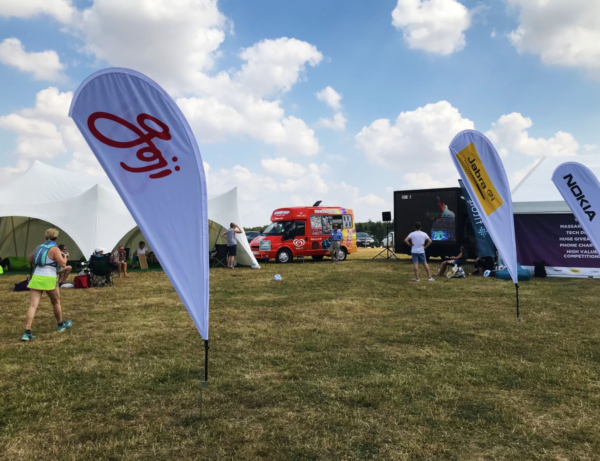 The Race to the Stones base camp with sponsor flags and a food truck on a sunny summer day, with a runner and spectators visible on the grass.