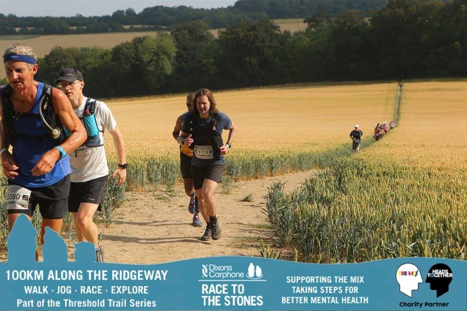 Three runners including bib 1113 descend a narrow path between golden wheat fields during the Race to the Stones, with a long line of participants stretching into the distance.