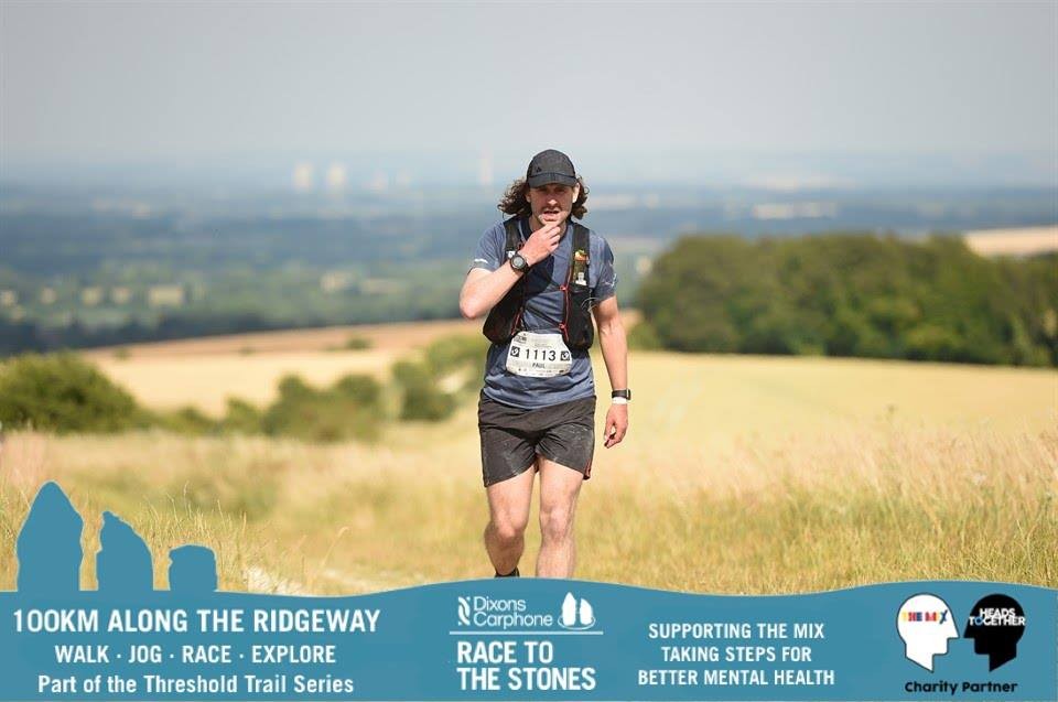 A male runner in a cap and race vest walks across open downland above a sweeping valley during Race to the Stones, the landscape stretching to the horizon behind him.
