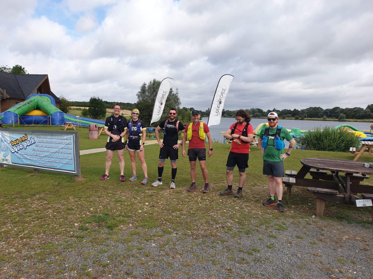 A group of six runners in race gear pose together at the lakeside start area of an ultra event, event banners and an inflatable water park visible behind them.