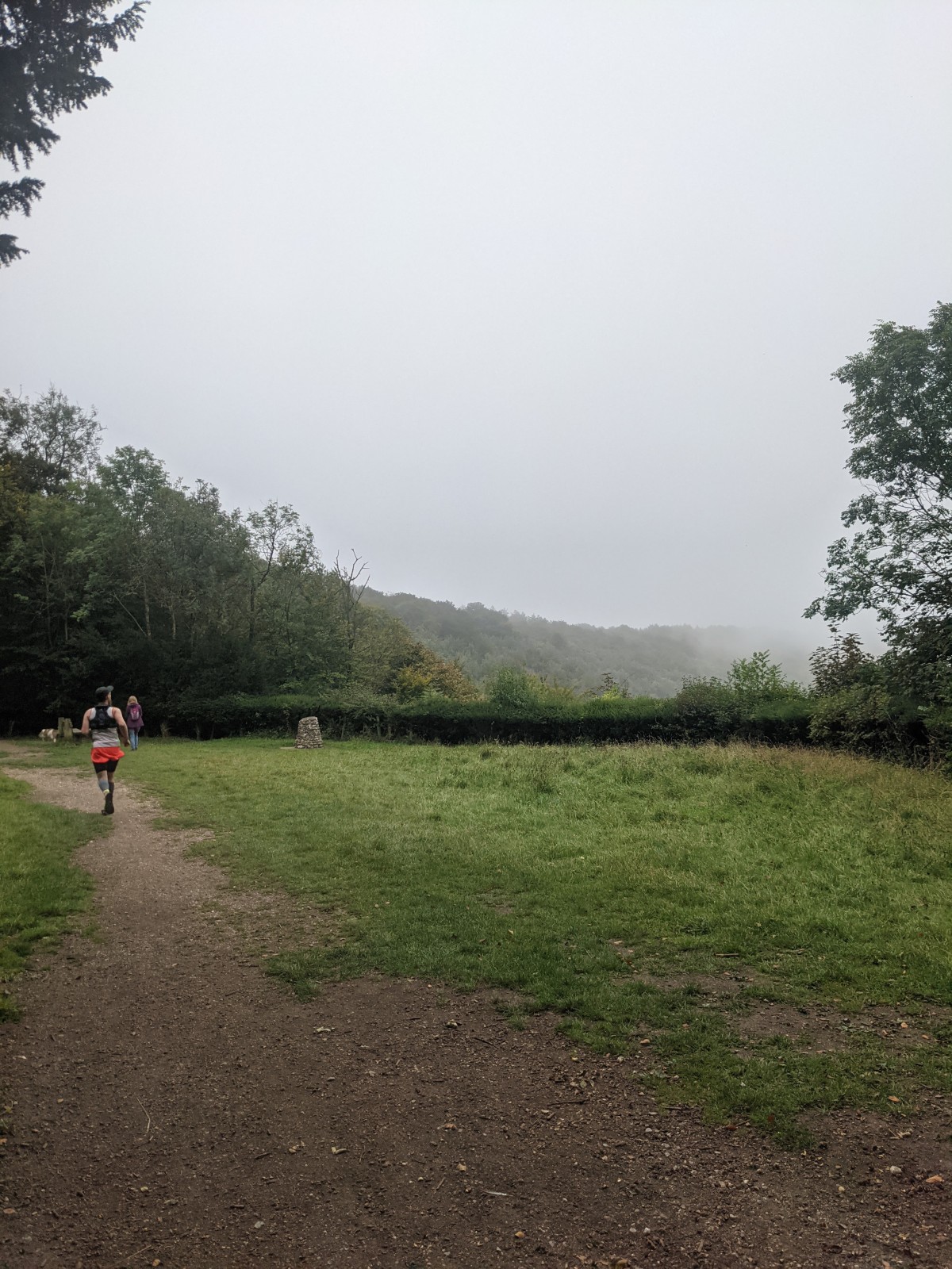 A runner in orange shorts heads away from the camera along a dirt path beside open misty hillside, a trig point visible in the middle distance.