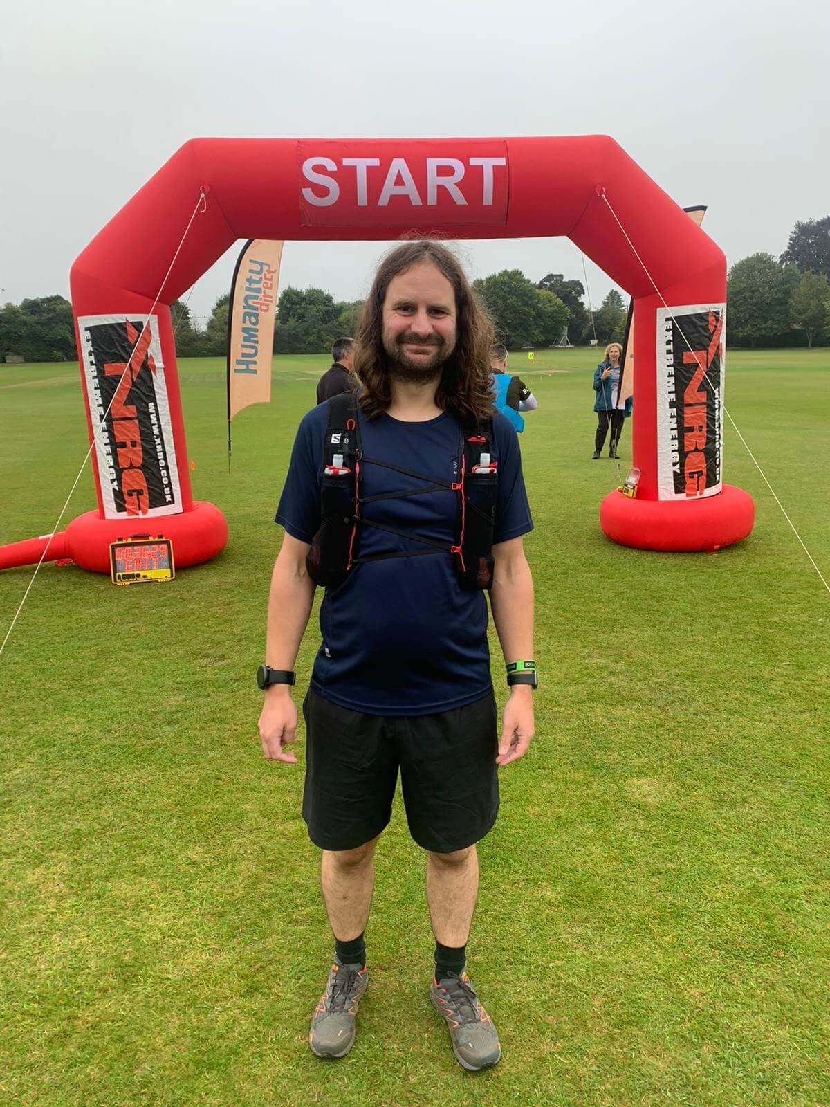 A male runner stands beneath the red START arch of the Humanity Direct ultra race on a cricket ground, wearing a hydration vest and looking relaxed before the race.