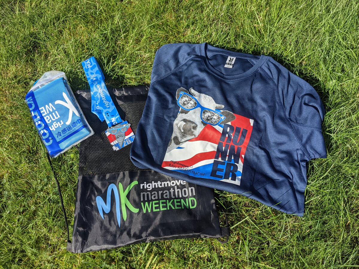 The MK Marathon Weekend goody bag laid out on grass, including a navy RUNNER t-shirt, a medal on a blue ribbon, and a drawstring bag.
