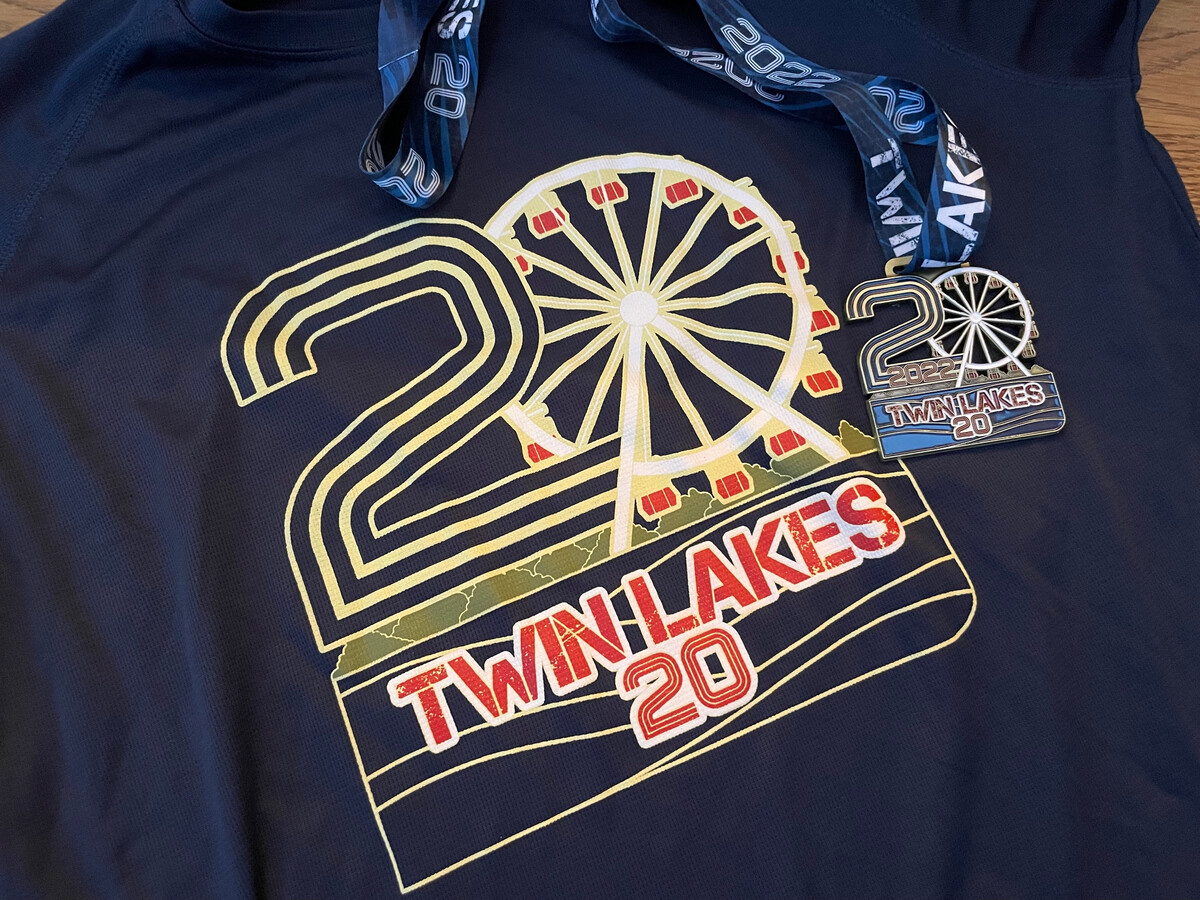 A Twin Lakes 20 race t-shirt and matching medal laid flat, the shirt showing a Ferris wheel design and "Twin Lakes 20" text in gold.