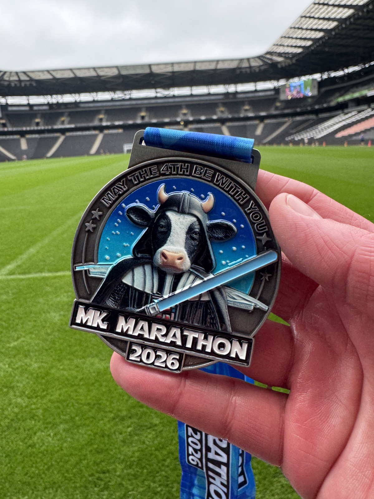A hand holds up the 2026 MK Marathon medal inside the stadium, a detailed cow dressed as Darth Vader design with the text "May the 4th Be With You" on a blue ribbon.